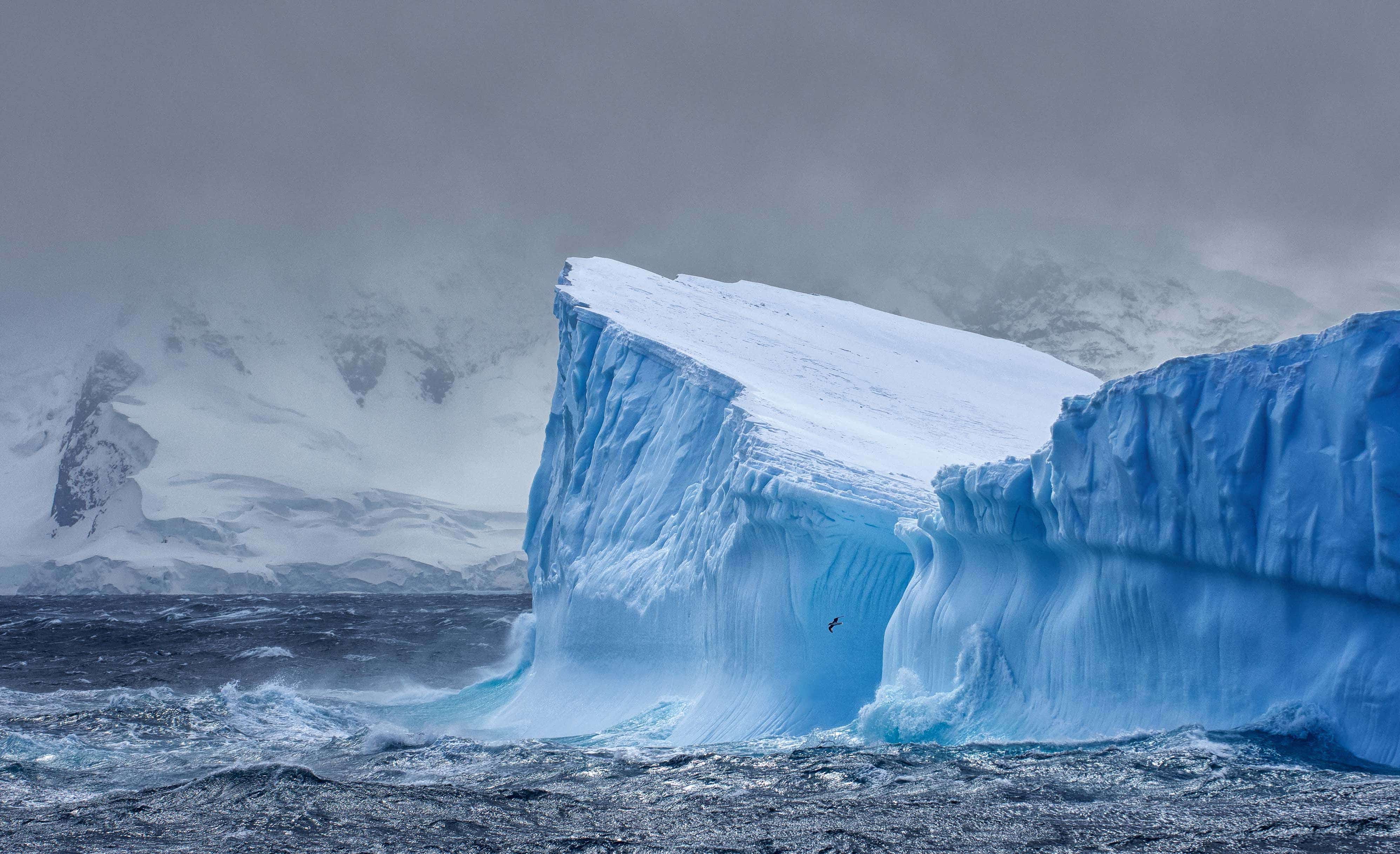 Et stort blåt isbjerg i havet, men et bjerg i baggrunden